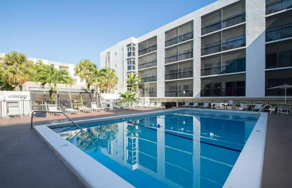 Original Real estate property photo: The photo depicts an outdoor pool area at a residential complex, bathed in bright natural light. While the image captures the setting well, there's an opportunity to enhance the warmth and saturation to make the scene more inviting, emphasizing the vividness of the pool and surrounding greenery.