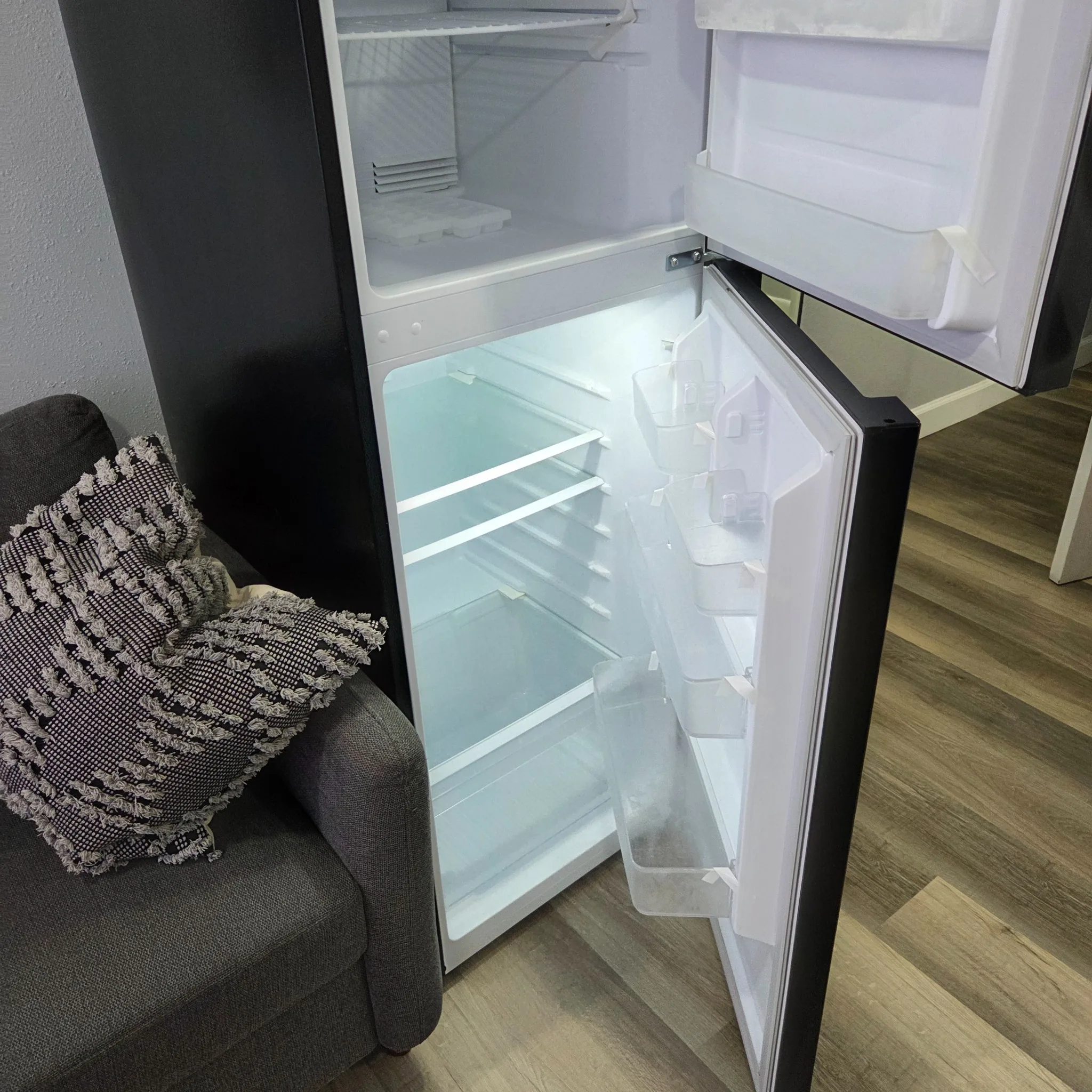 Original Open refrigerator next to a sofa with a decorative pillow on a room with wooden floor.