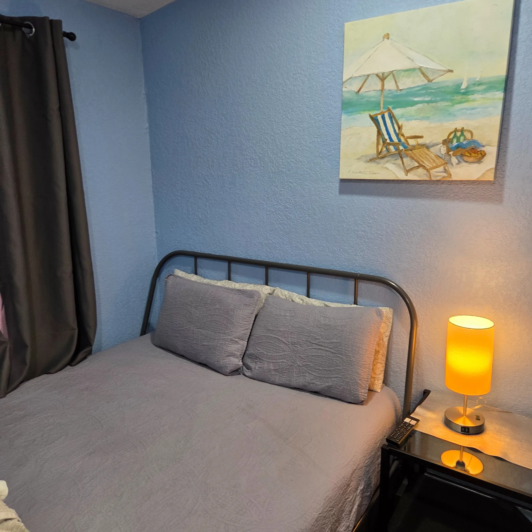 Original Cozy bedroom with a metal bed frame, soft bedding, a painting, and a lit bedside lamp on a table.
