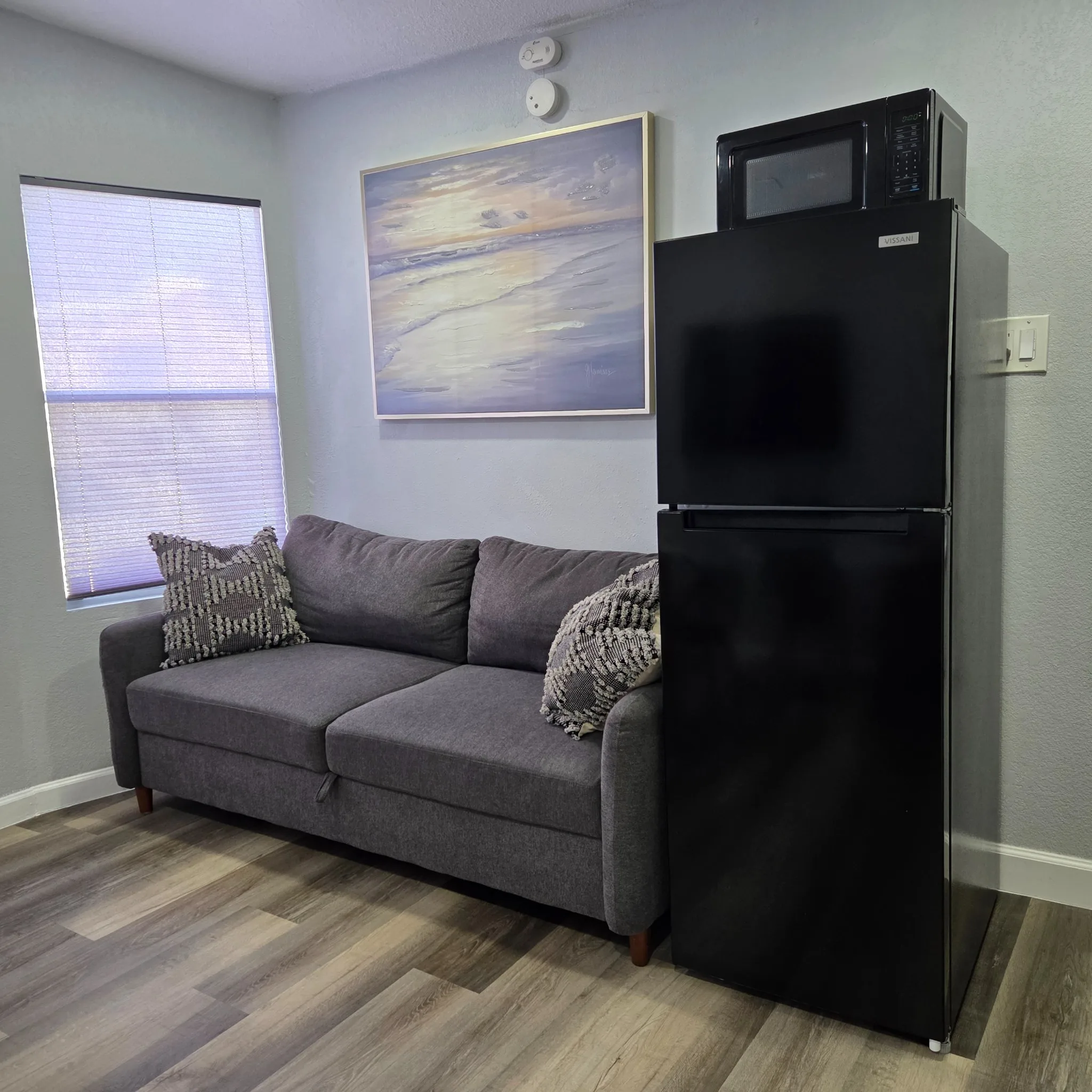 Original Small living area with a gray sofa, decorative pillows, a fridge, and wall art under natural light from a window.