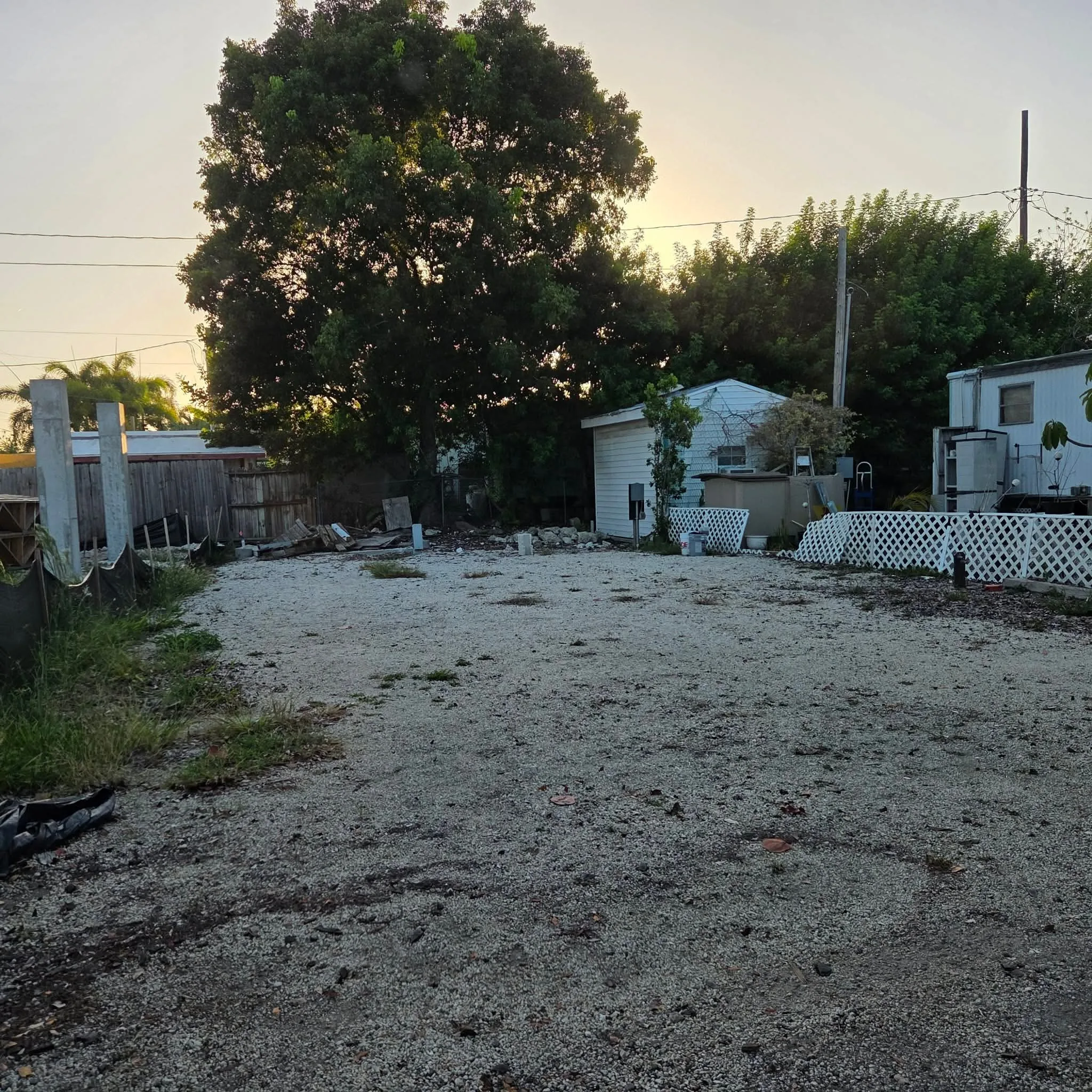 Original Outdoor gravel area with surrounding trees, a small structure, and fencing, captured at dusk with muted lighting.