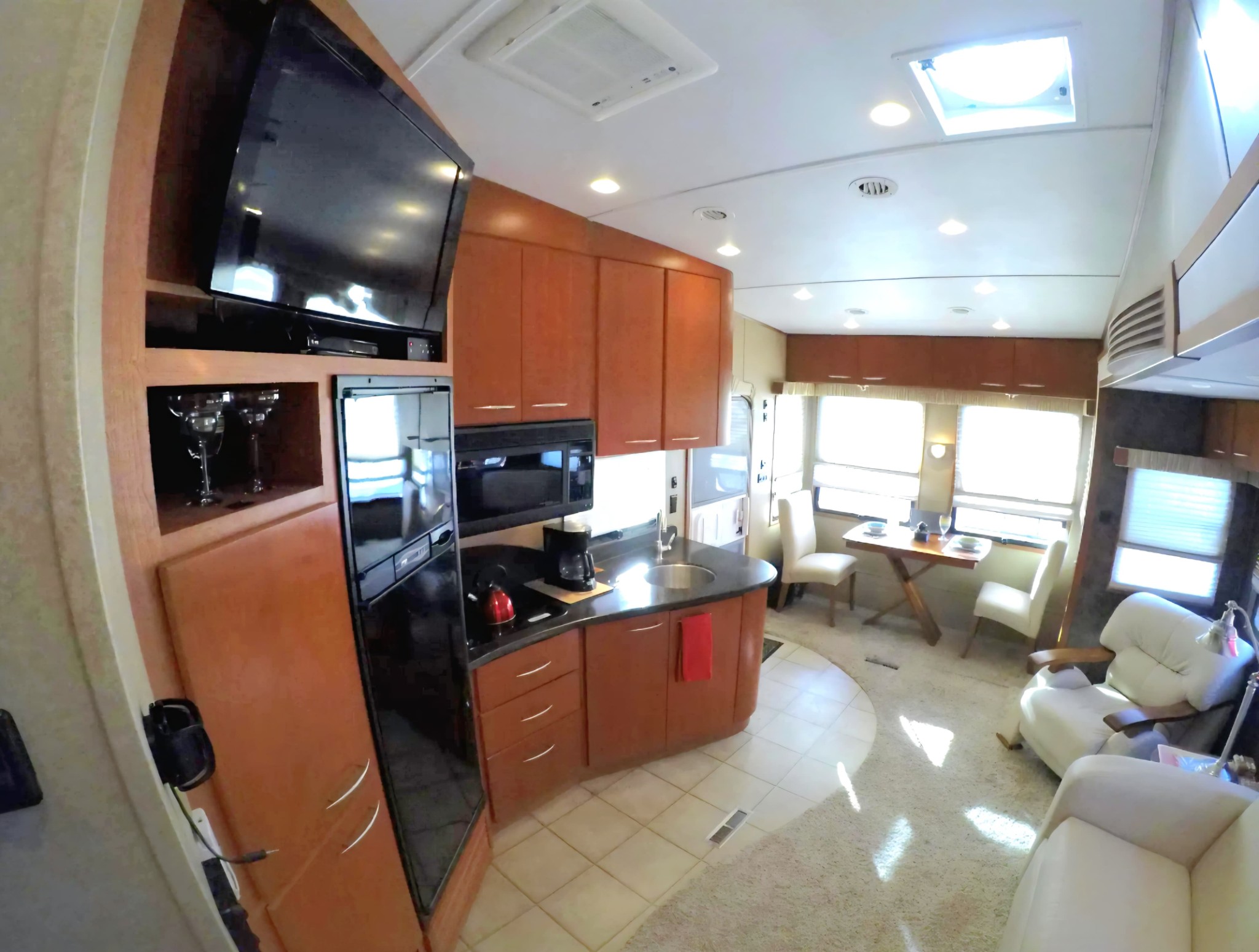 Original Real estate property photo: The photo captures a compact kitchen and dining area within a recreational vehicle, featuring warm wood cabinetry and ample natural light. The image appears slightly overexposed with some areas lacking detail due to strong sunlight. To ensure MLS compliance, adjustments will focus on balancing brightness and contrast to enhance clarity while maintaining a natural appearance.