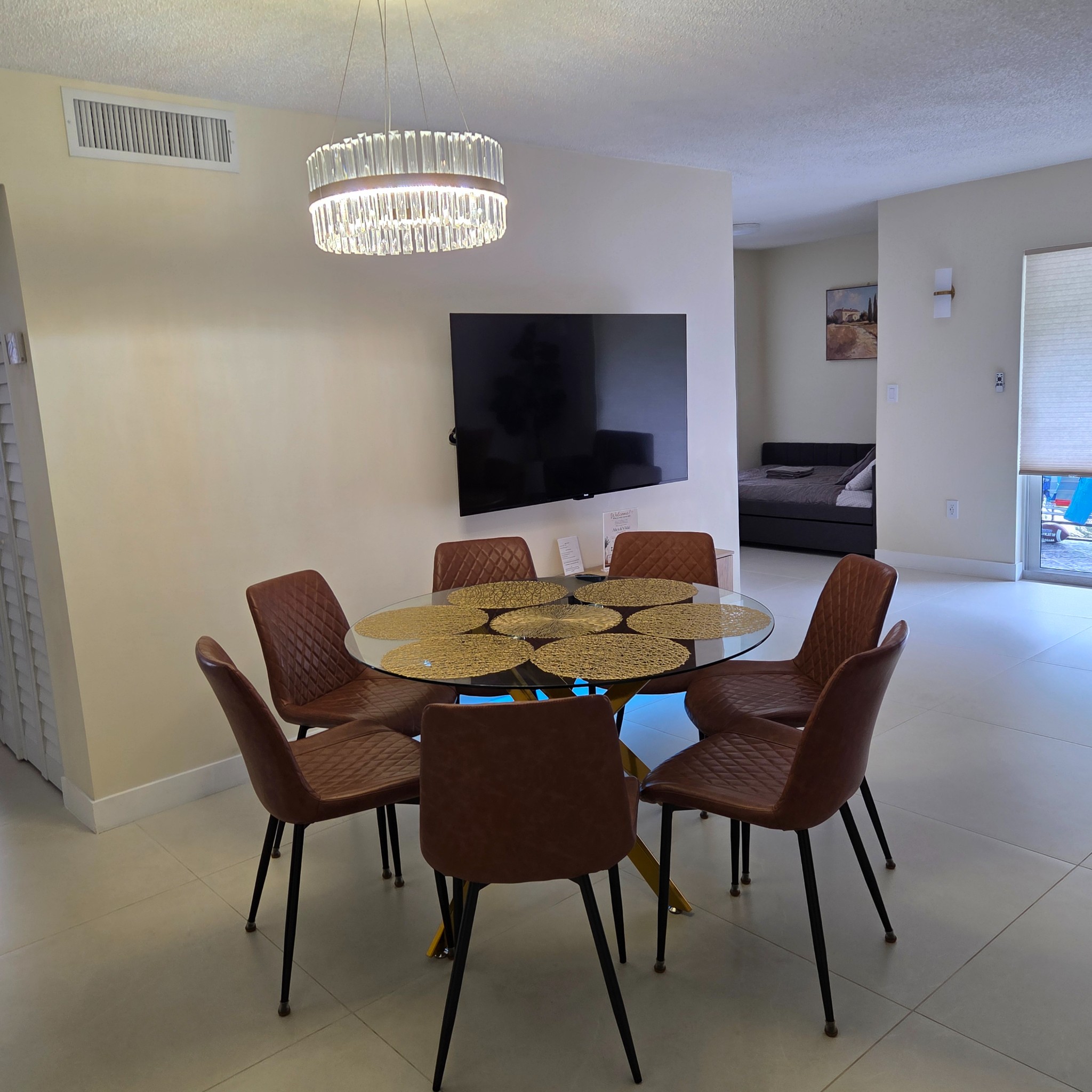 Original Real estate property photo: The photo depicts a dining area with modern furnishings and a large wall-mounted TV. The lighting appears slightly dim with a cool cast, which can be enhanced for clarity and warmth. To achieve a natural and inviting look, adjustments to brightness and warmth will help balance the overall tone.