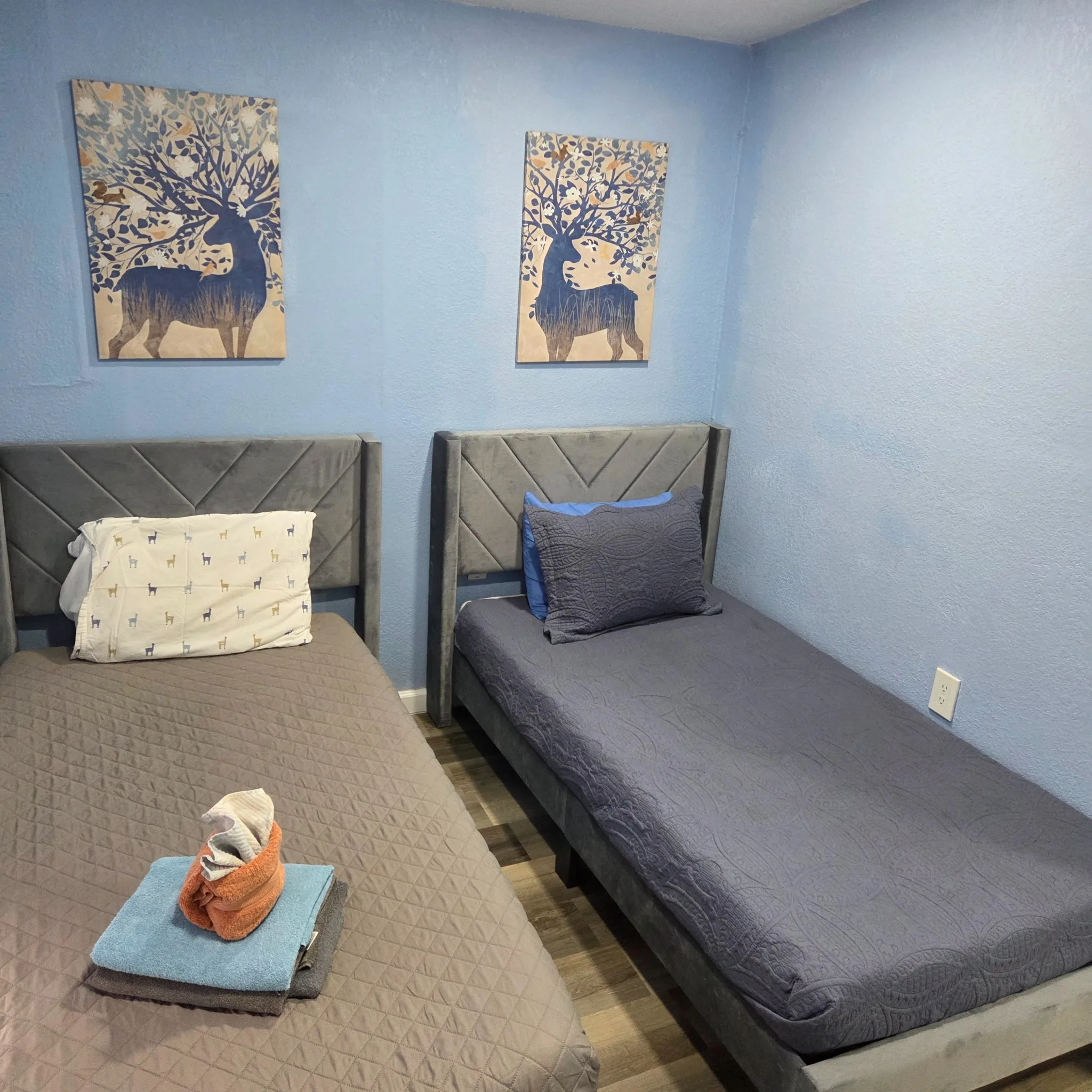 Original Small bedroom with twin beds, blue walls, and decorative art featuring deer. The beds have matching gray and blue bedding.