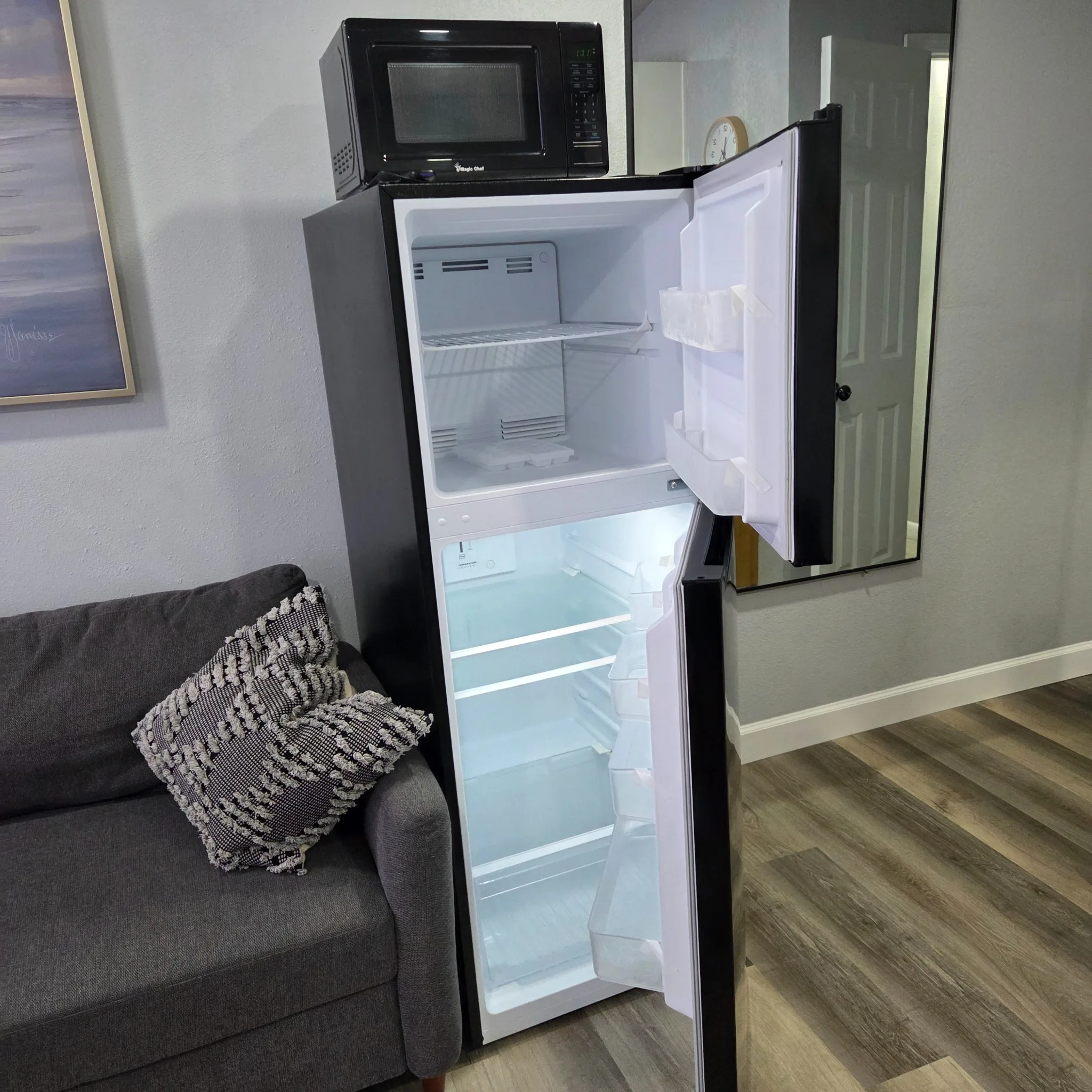 Original Living area with a dark gray couch, a pillow, and an open refrigerator under a microwave, featuring a wall clock and artwork.