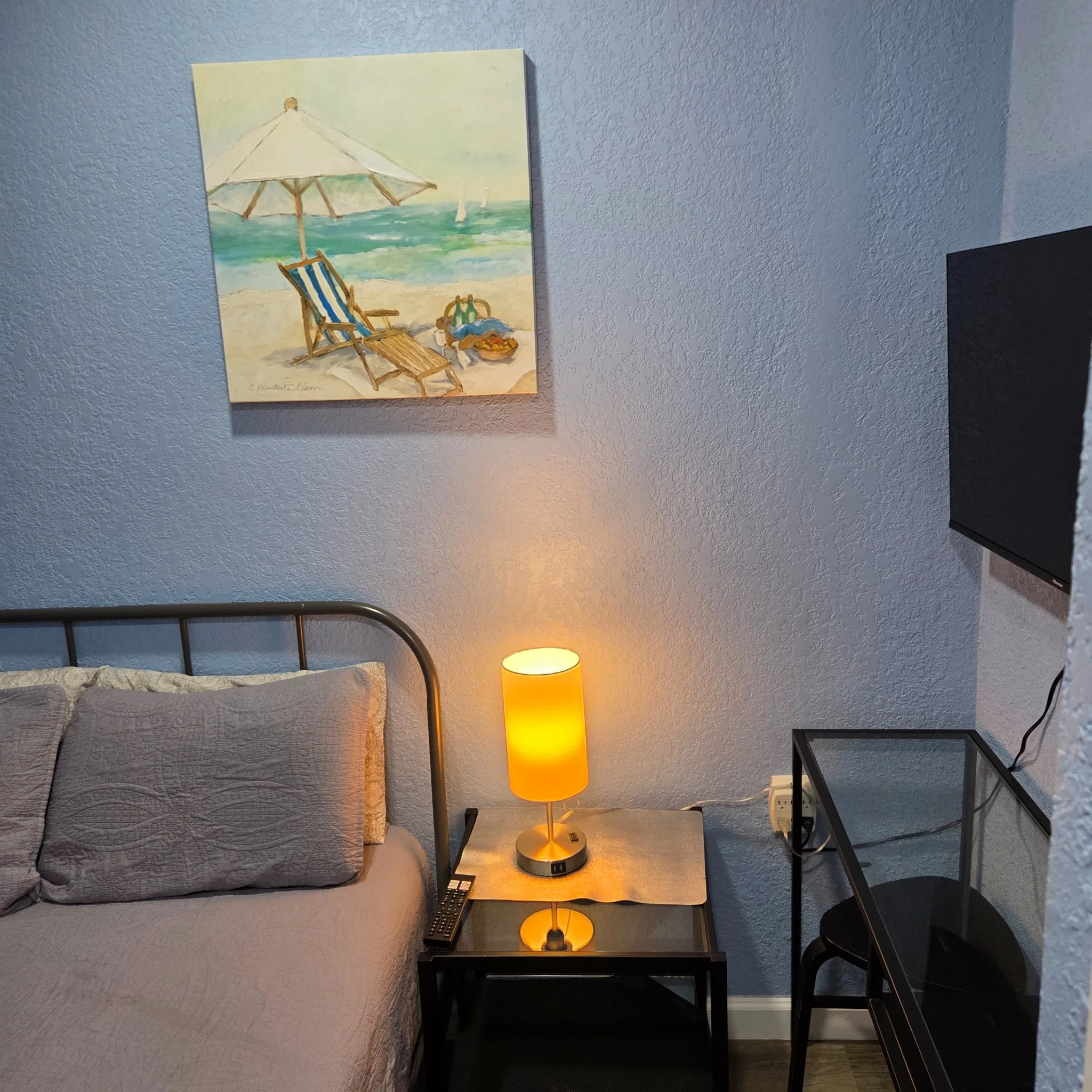 Original Bedroom corner with a bed, bedside table, lamp, beach painting, and a wall-mounted TV.