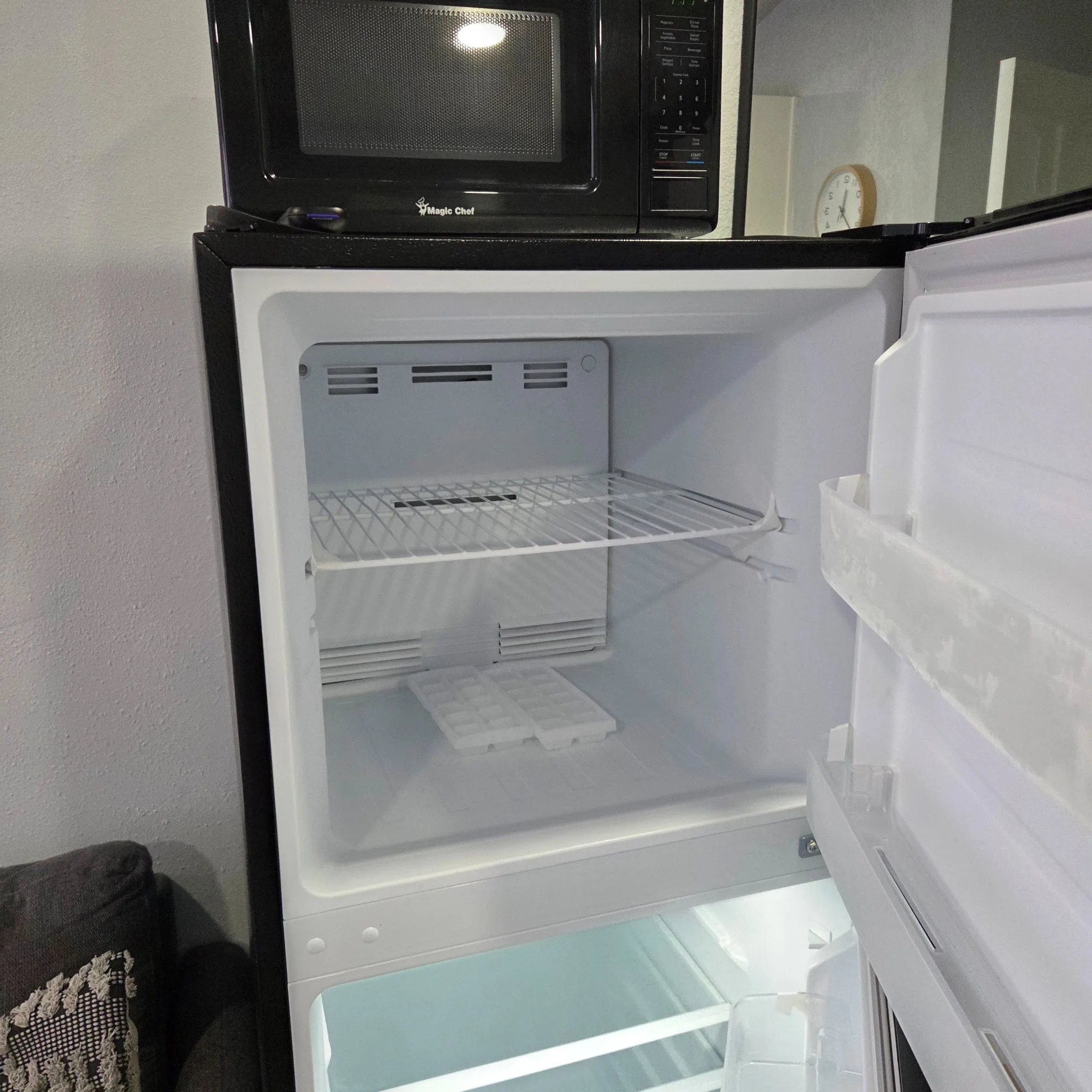 Original Open refrigerator with empty shelves and ice tray, topped by a black microwave in a kitchen setting.