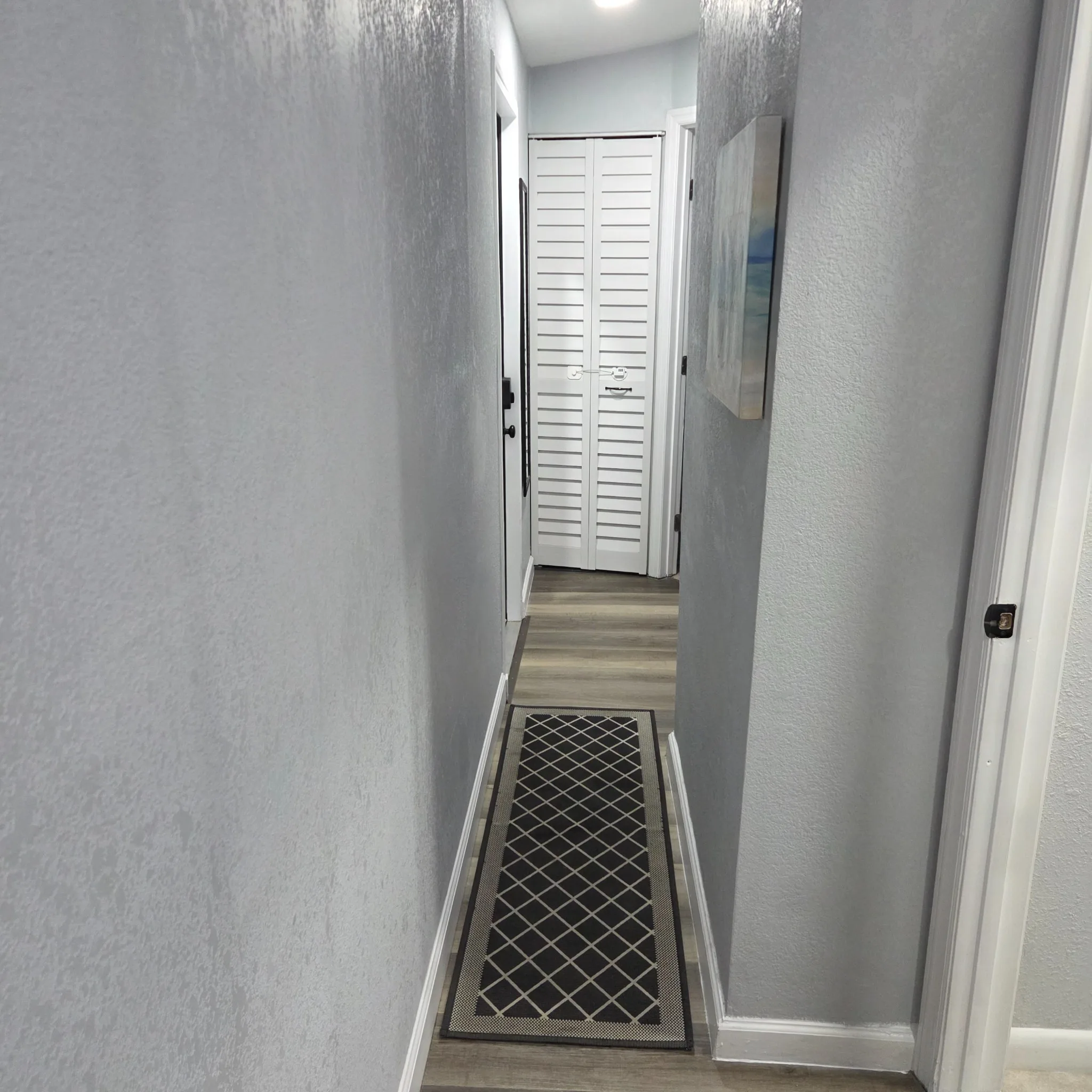 Original A narrow hallway with light gray walls, wooden flooring, and a decorative rug leading to a door with white shutters.