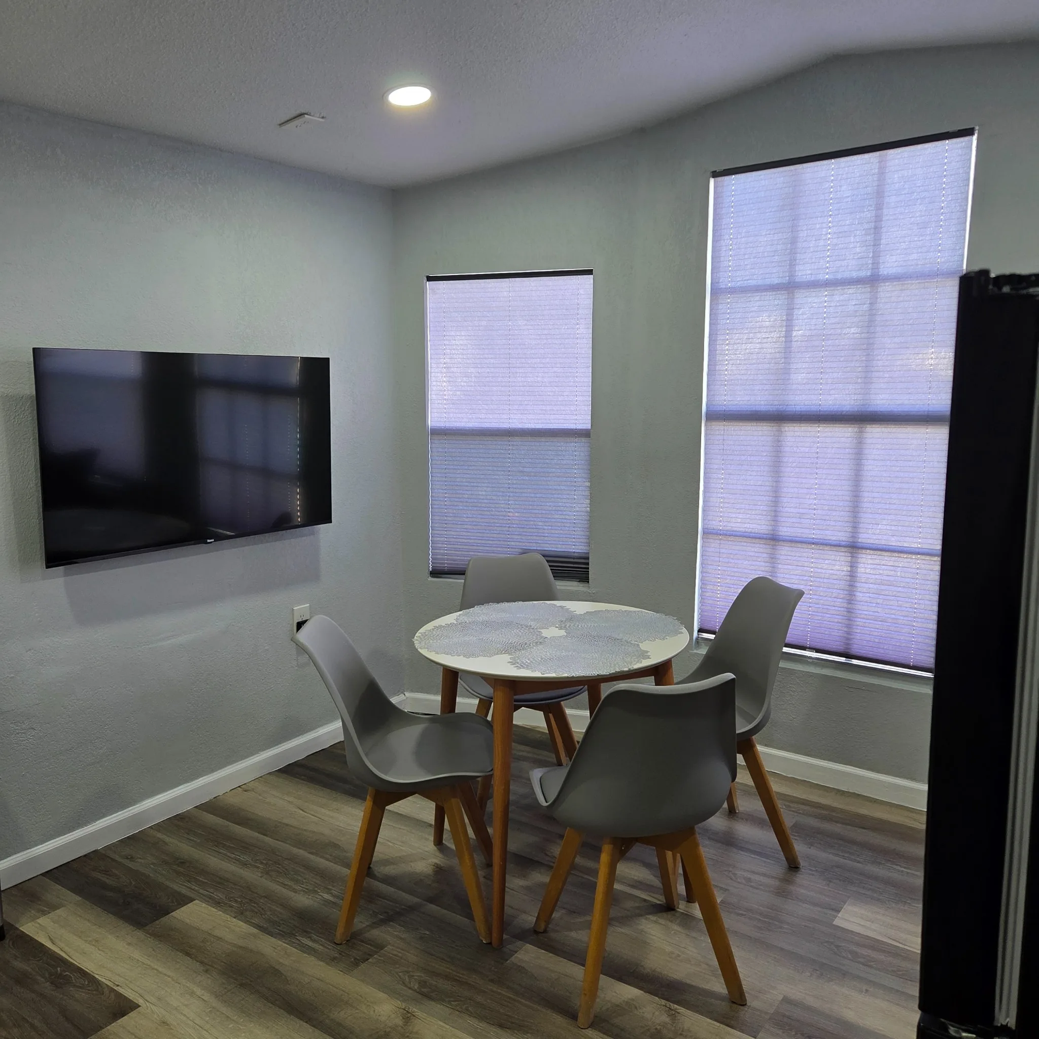 Original Dining nook with gray chairs, round table, wall-mounted TV, and large windows with shades.