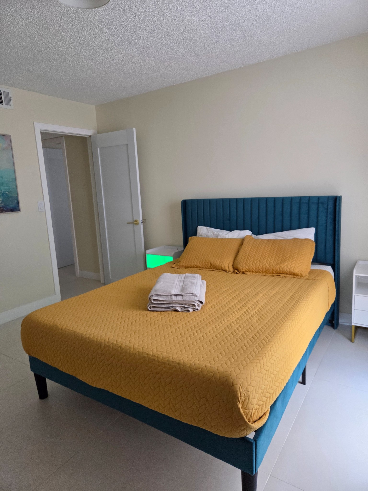 Original Real estate property photo: The bedroom photo features a yellow bedspread on a blue bed against light-colored walls. The image suffers from slightly dull lighting, particularly in the corners, and has a cool color cast. Enhancements will focus on improving brightness and contrast to highlight details while maintaining a realistic look in line with MLS requirements.